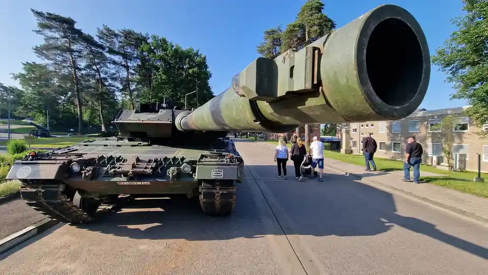 A large green military tank with an extremely long cannon barrel is parked on a paved road, its barrel angled upward…