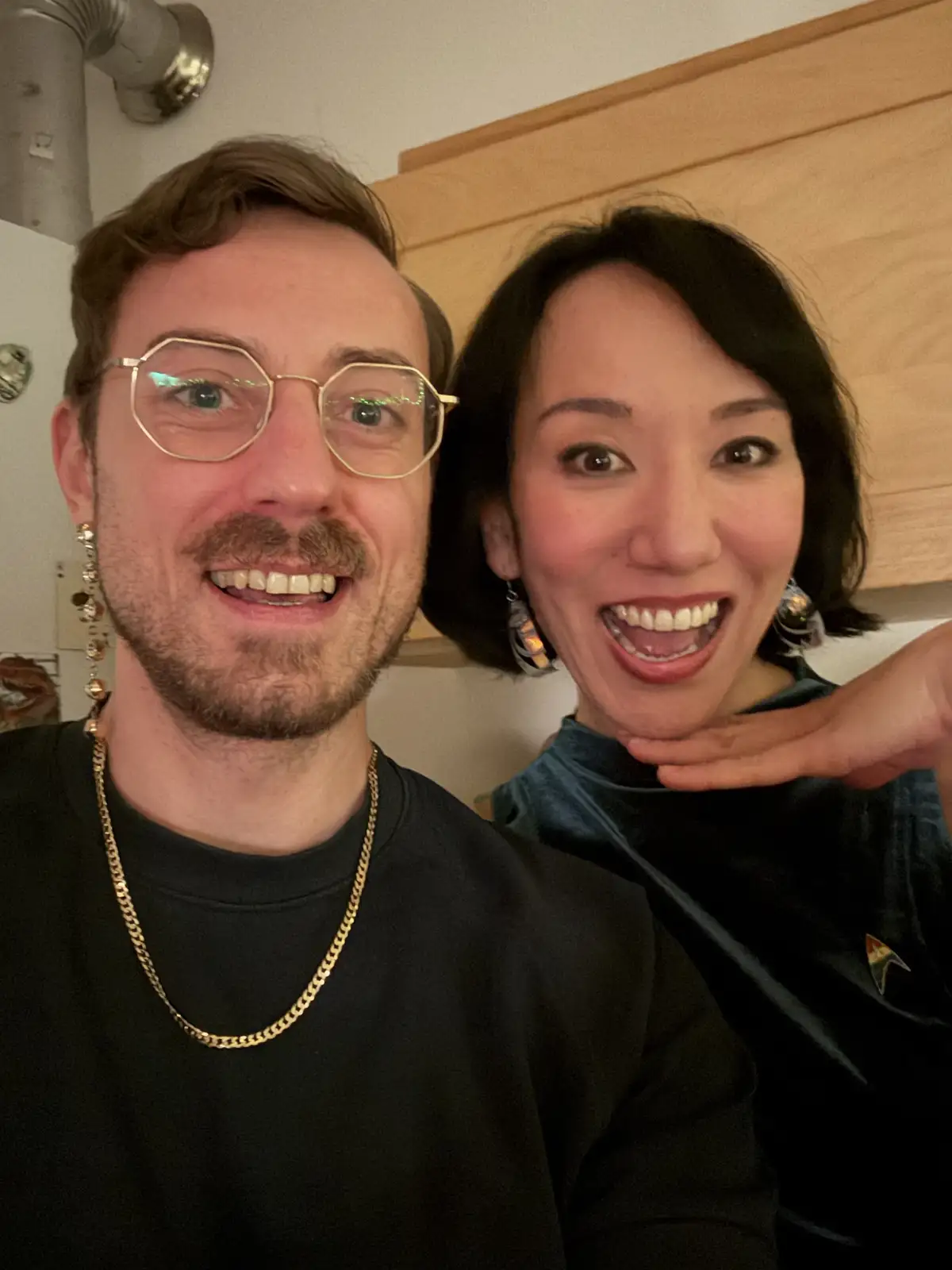A selfie of a man and a woman smiling at the camera in a kitchen. The man wears gold-rimmed glasses, a black shirt, and a…