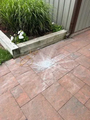 A wet, circular patch on a red brick patio floor resembles a snowflake, with a yellow garden hose coiled nearby and a wooden…