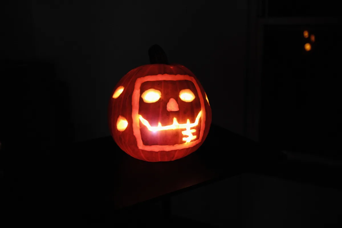 A carved jack-o'-lantern with a glowing face sits in a dark room, its eyes, nose, and jagged mouth illuminated from within…