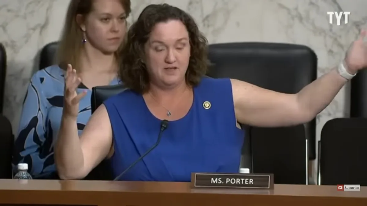 A woman in a blue sleeveless top sits at a wooden table with a nameplate reading 'MS. PORTER' and a microphone, gesturing…