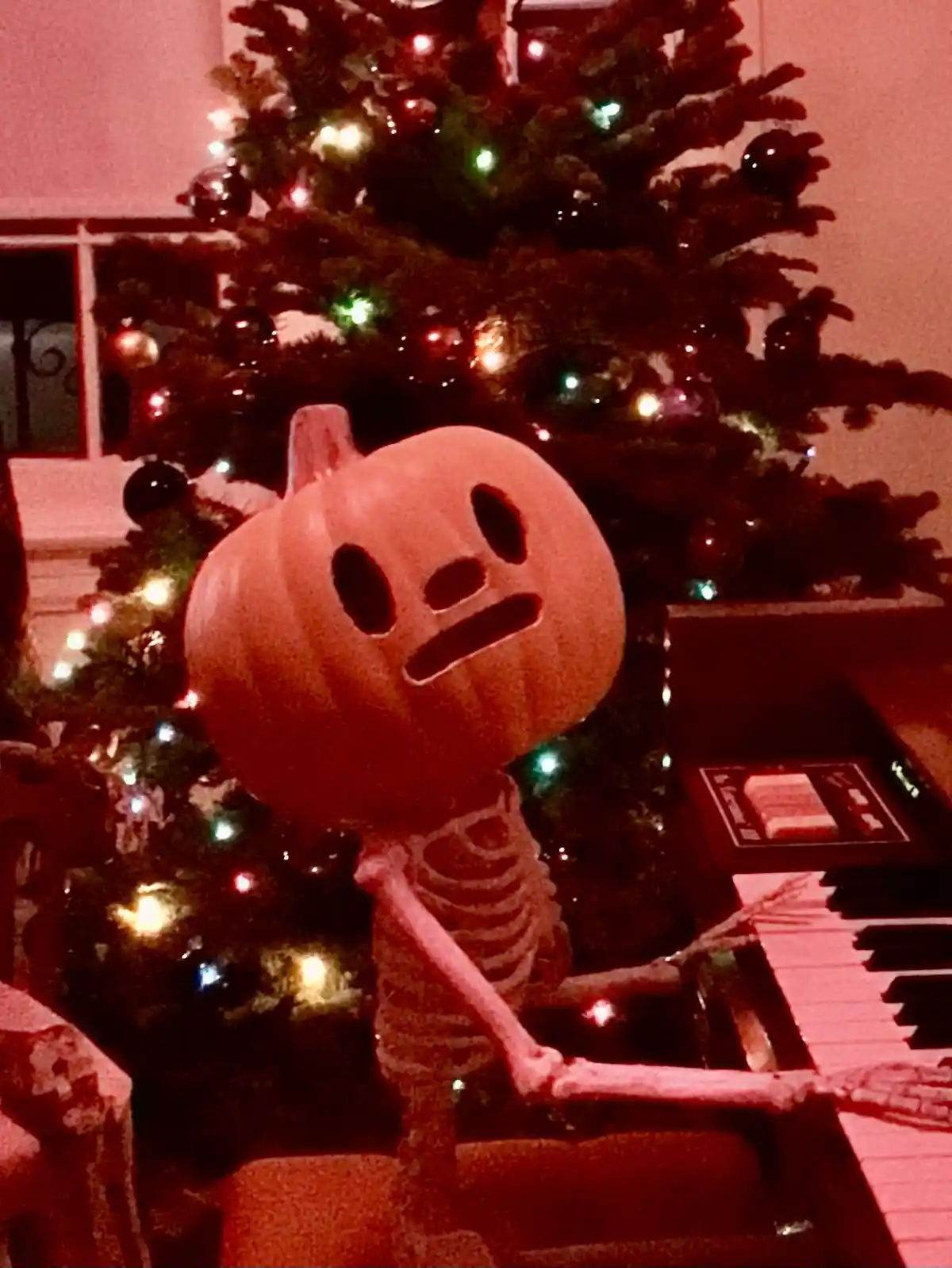 A skeleton with a carved pumpkin head sits at a piano in front of a lit Christmas tree, looking confused or annoyed. The…