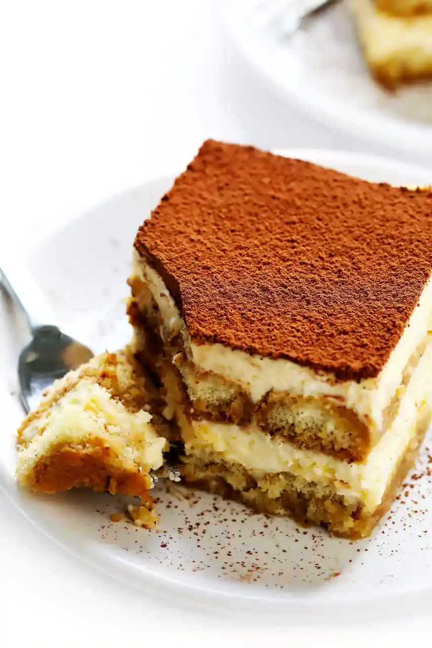 A close-up shot of a square slice of tiramisu on a white plate, dusted with cocoa powder, with a bite taken out and a spoon…