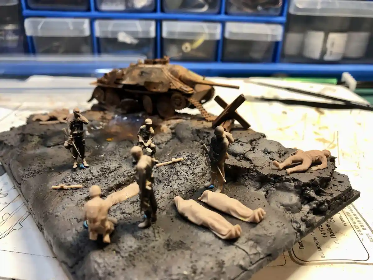 A diorama of miniature World War II soldiers and a tank on a muddy, textured base, with several figures posed as if in…