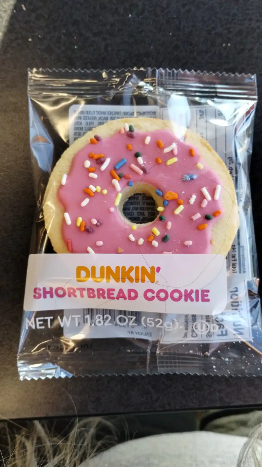 A close-up of a Dunkin' branded shortbread cookie in its clear plastic packaging, shaped like a donut with pink frosting and…