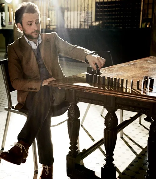 a man in a brown suit sits at a wooden table, carefully placing a domino tile in a line of standing dominoes. The scene is…