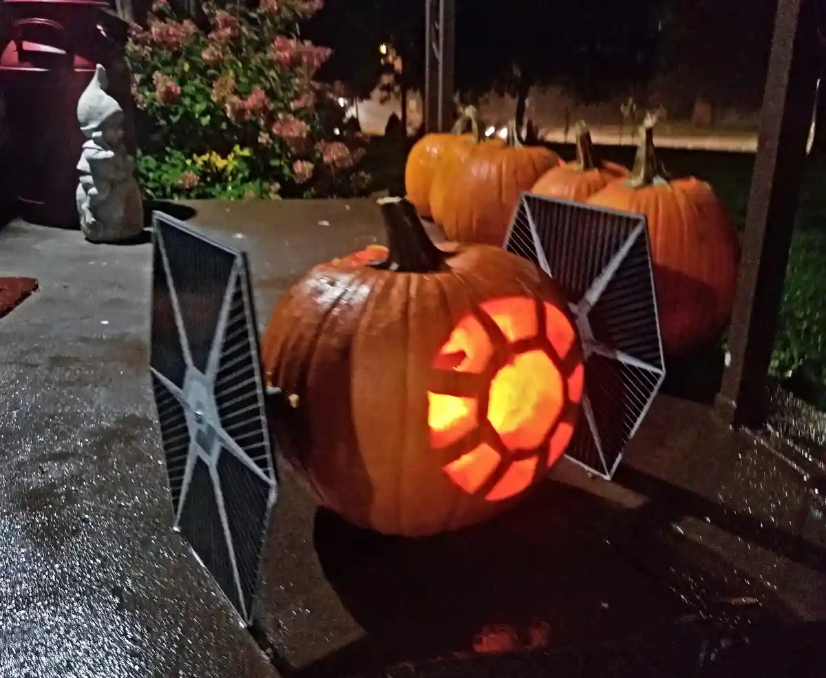 A carved Halloween pumpkin with a glowing interior sits on a wet porch, flanked by two small, solar-powered TIE fighter…