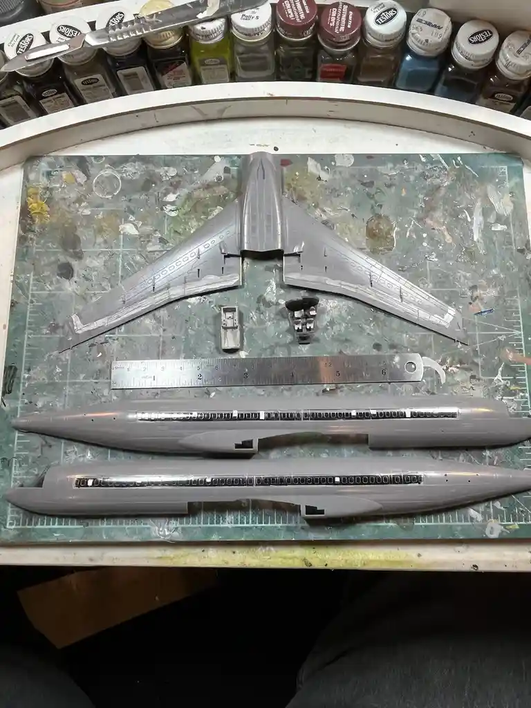 A top-down photo shows unassembled grey plastic parts of a model airplane, including wings and fuselage halves, laid out on…