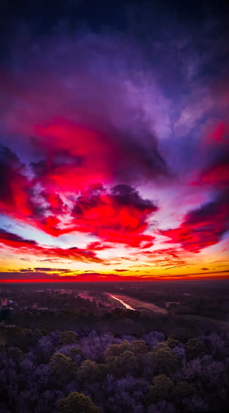 A vibrant, high-angle aerial photograph captures a dramatic sunset or sunrise over a forested landscape, with clouds painted…