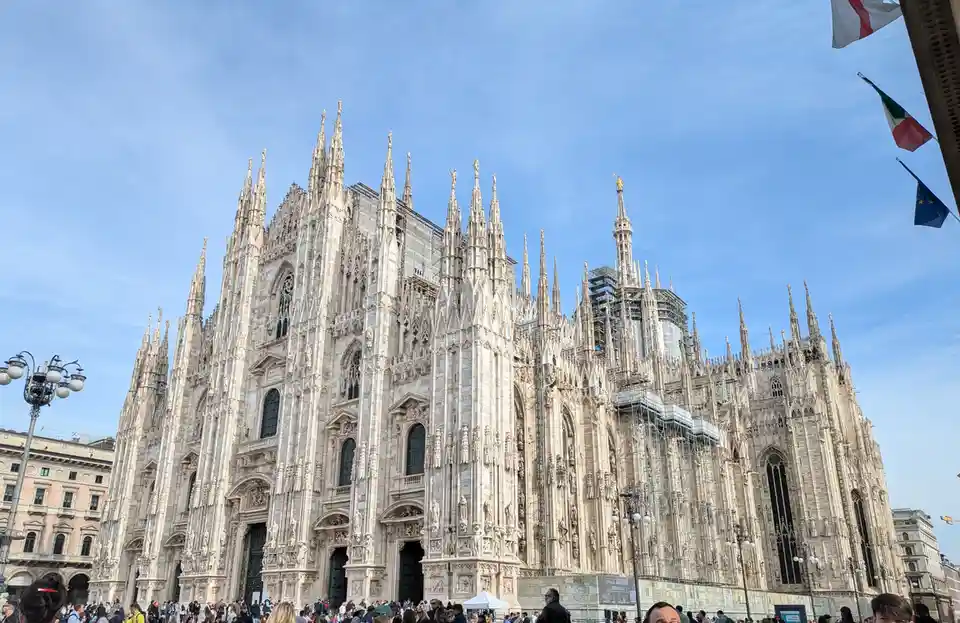 The image shows the ornate, Gothic-style Milan Cathedral (Duomo di Milano) under a clear blue sky, with extensive…