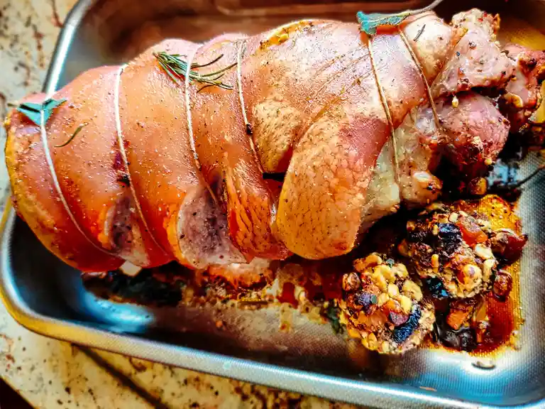 A close-up photo of a roasted pork roast in a metal baking tray, tied with twine and garnished with rosemary and bay leaves…