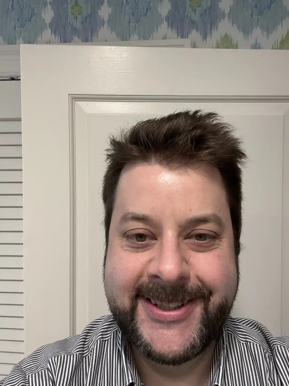 A man with a beard and short brown hair smiles at the camera while wearing a black-and-white striped shirt, standing in…