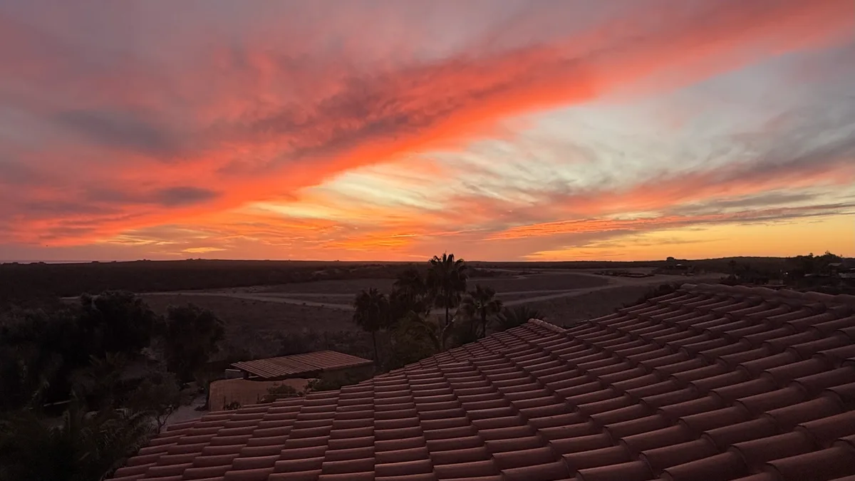 A scenic photo of a vibrant orange and pink sunset over a desert landscape, viewed from atop a terracotta-tiled roof with…