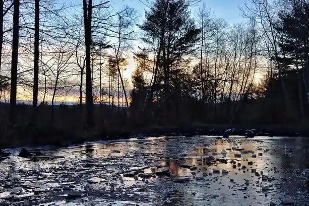 A serene, low-light photograph of a river at dusk or dawn, with broken ice floes floating on the water's surface. Bare trees…
