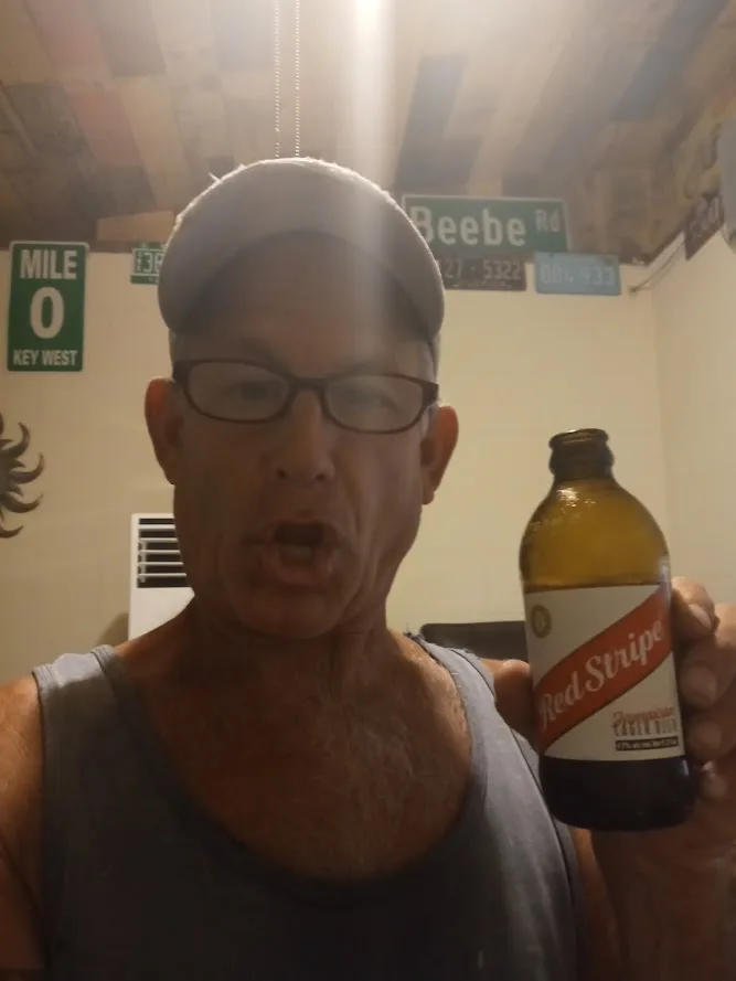 A man wearing glasses and a cap holds up a bottle of Red Stripe beer while making a surprised or shocked facial expression…