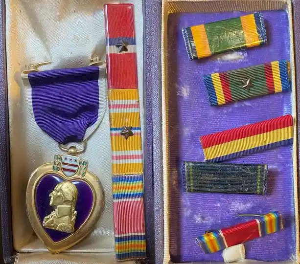 A close-up photo shows a military medal case open, displaying a Purple Heart medal on the left with its purple ribbon and…