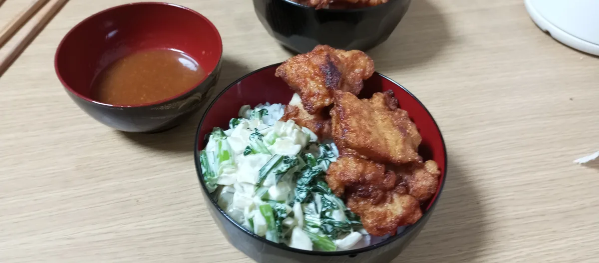 A close-up photo shows a black bowl with a red interior containing fried chicken pieces, white rice, and a creamy green…