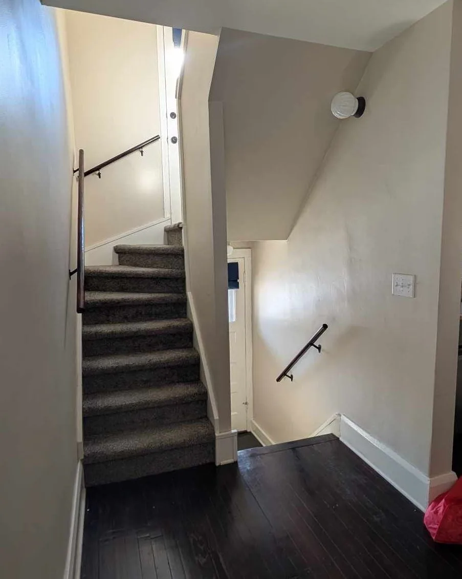 A photo of a narrow staircase with carpeted steps and wooden handrails, leading upward from a dark hardwood floor. The walls…