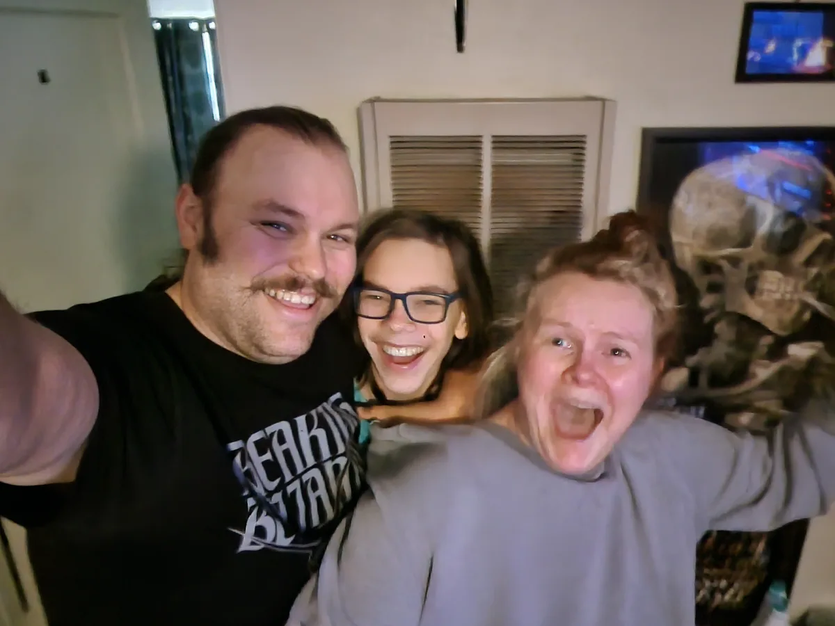 Three people are taking a selfie in a living room, with a large skull decoration visible behind them. The person on the left…