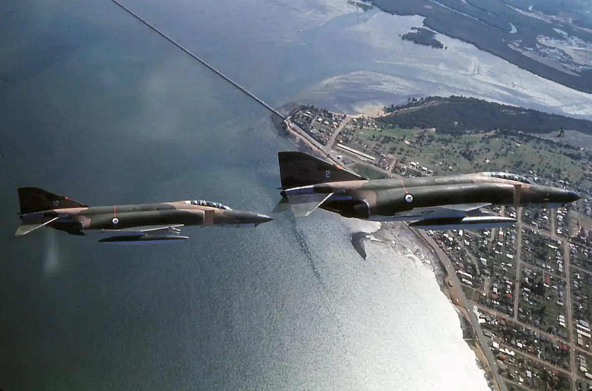 Two military jets fly in formation over water and land, connected by a refueling hose from the right jet to the left. The…
