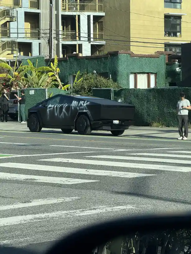 A black, angular Tesla Cybertruck is parked on a city street with white crosswalk lines visible in the foreground. The side…