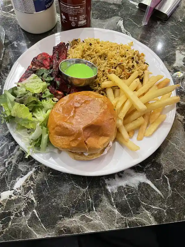 A white plate holds a burger, fries, fried rice, grilled meat, and lettuce on a dark marble table. A small metal cup of…