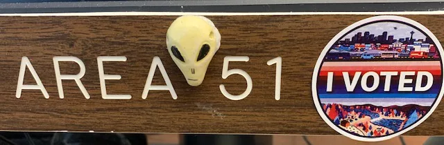 A close-up of a wooden sign reading 'AREA 51' with a small alien head figurine glued above the '5'. To the right is a…
