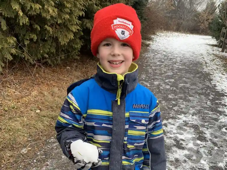 A young boy in a red 'Scotiabank Hockey Club' beanie and a blue striped winter jacket stands outdoors on a snowy path…
