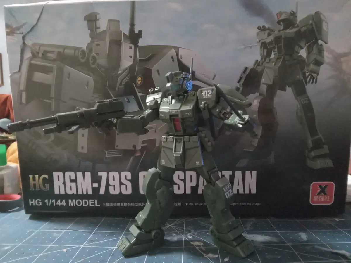 A green, assembled 1/144 scale model of the RGM-79S Spartan mobile suit stands in front of its original packaging box. The…