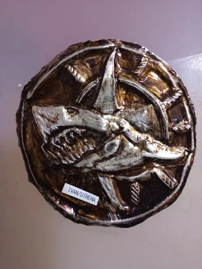 A circular, metallic-looking artwork depicting a shark with an open mouth and visible teeth, rendered in a textured…