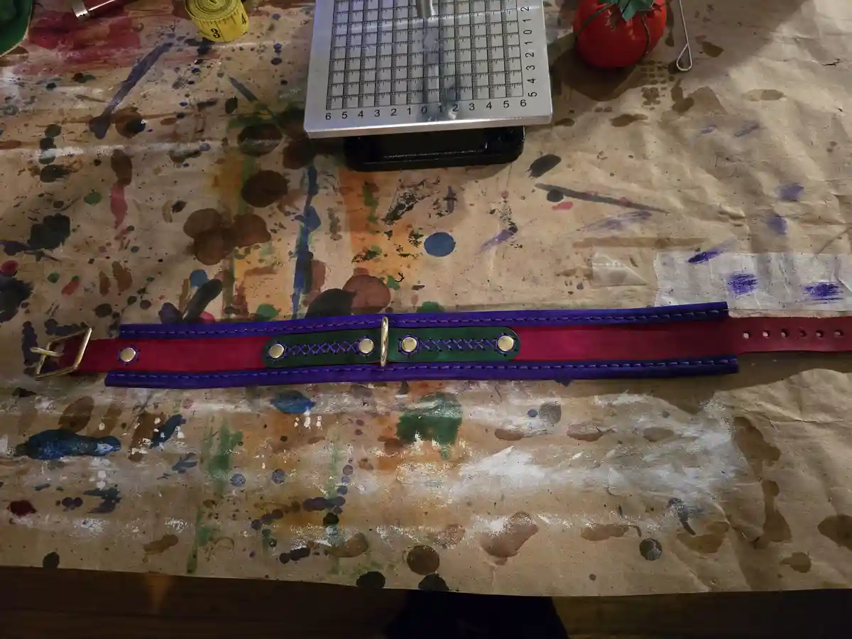 A handmade leather belt with purple and red straps, green central panel, gold hardware, and purple stitching lies on a…
