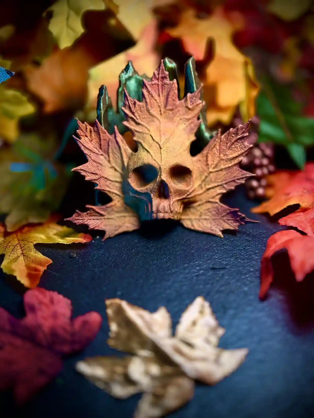 A stylized skull crafted from a maple leaf sits atop a bed of colorful autumn leaves on a dark surface, with the text…
