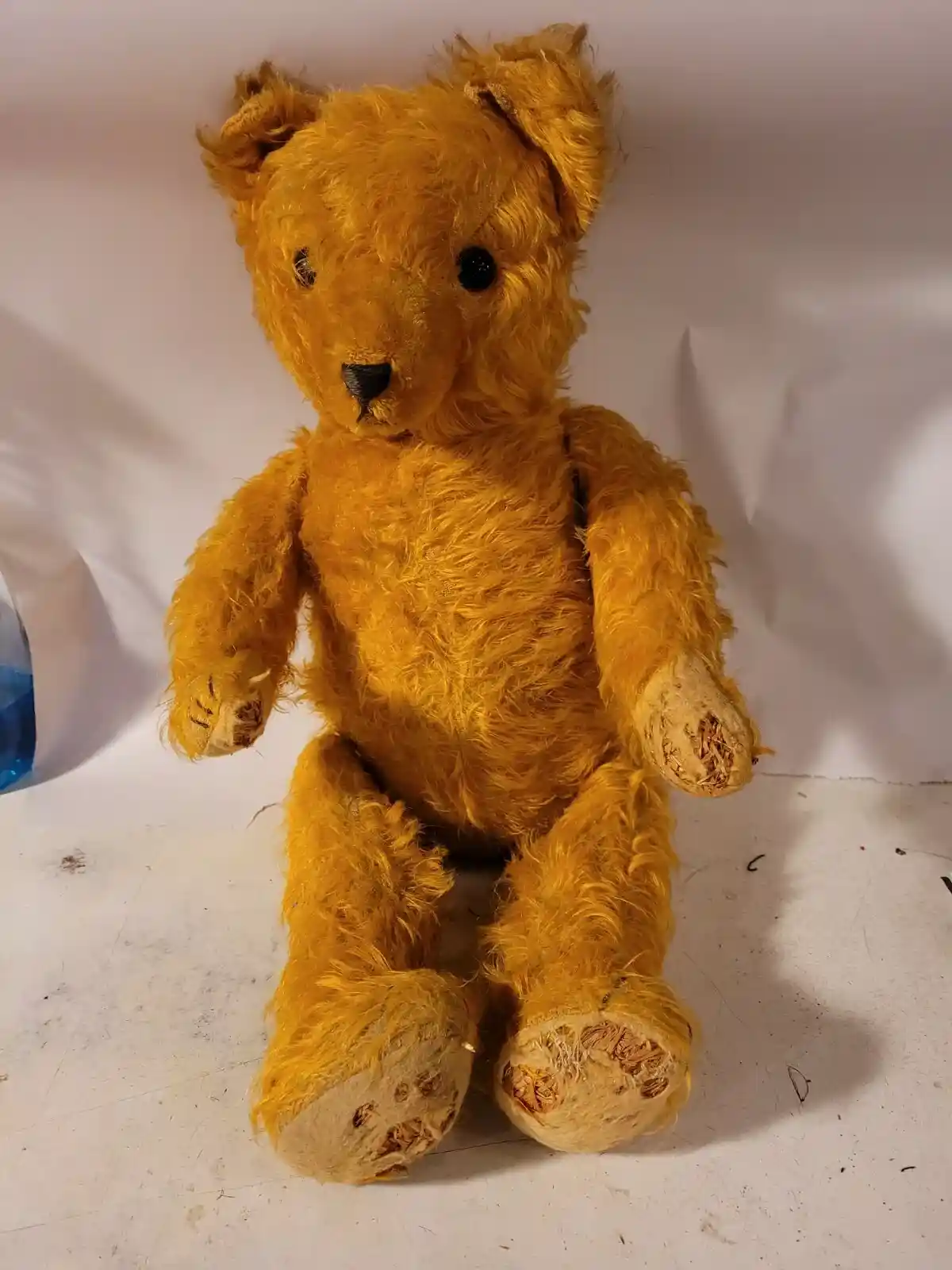 A very old, worn, golden-brown teddy bear with frayed fur and exposed stuffing in its paws and feet sits on a white surface…