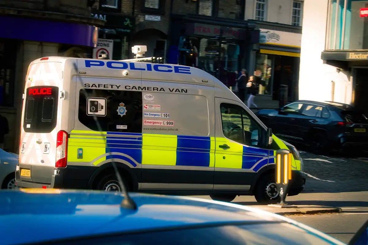 A white police van with blue and yellow markings is parked on a city street, labeled 'SAFETY CAMERA VAN' on its side. The…