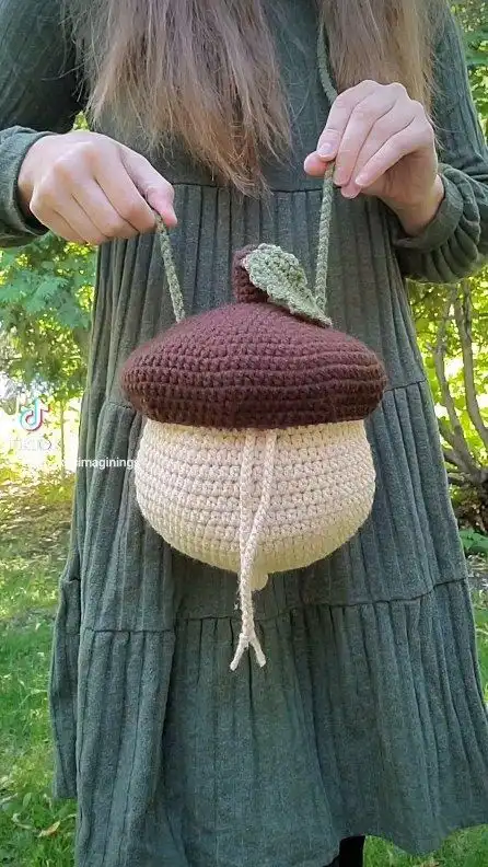 A person with long brown hair, wearing a green dress, holds up a handmade crocheted purse shaped like an acorn. The purse…