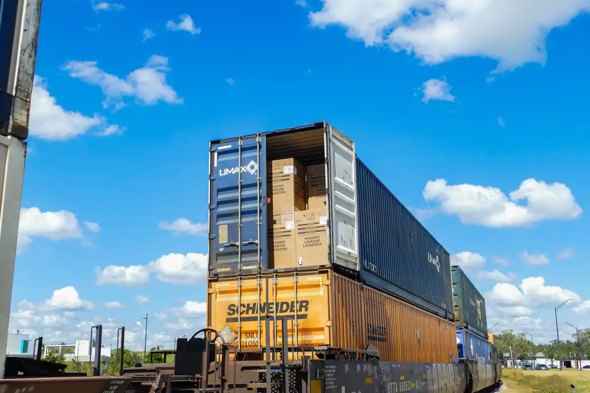 A freight train with stacked shipping containers under a bright blue sky with white clouds. One blue container is open…