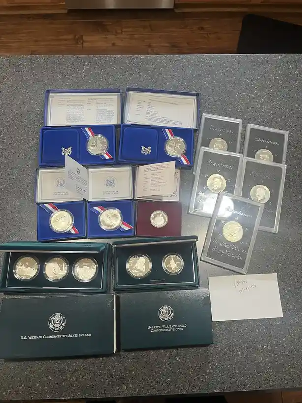 An overhead shot displays a collection of U.S. commemorative silver coins arranged on a gray countertop. Several coins are…