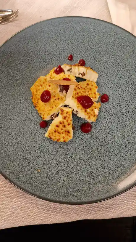 A top-down photo shows a grey, textured plate holding a dessert that looks like broken, golden-brown cheese pieces with red…