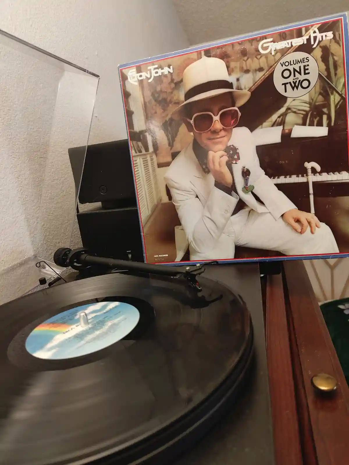 A vinyl record player has a record spinning on its platter, and the album cover for Elton John's 'Greatest Hits Volumes One…