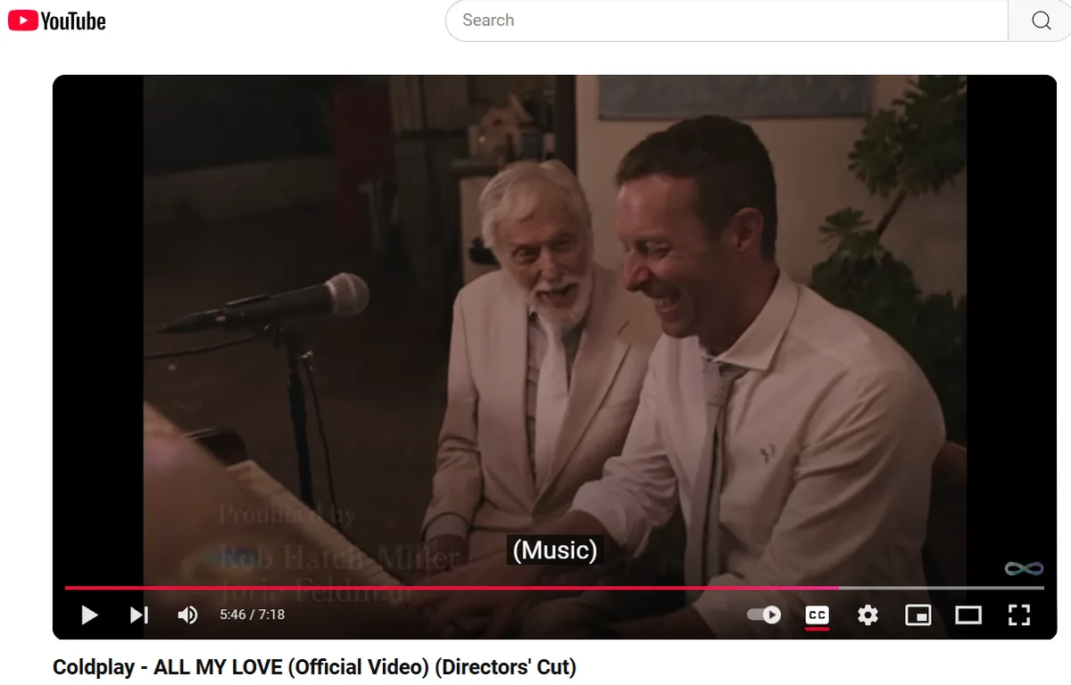 Two men in light-colored suits sit side-by-side, both laughing heartily while looking at something off-screen. A microphone…
