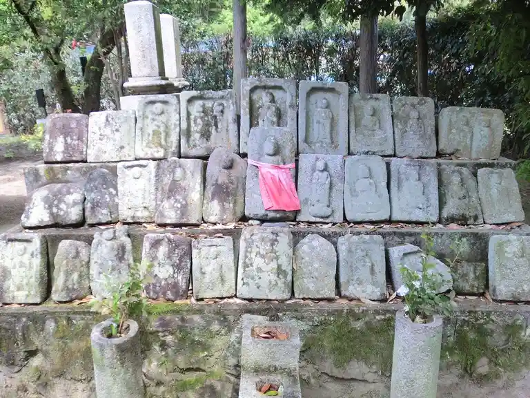 A stack of weathered stone tablets with carved figures, arranged in rows outdoors among trees and moss. One tablet in the…