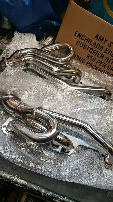 Two shiny chrome exhaust manifolds rest on bubble wrap, with a cardboard box behind them that reads 'AMY'S ENCHILADA BROTHER…