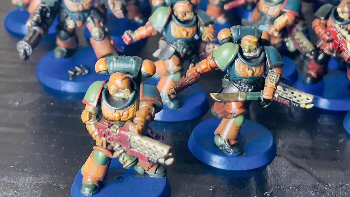 A close-up photo shows several painted miniature figurines of space marines in orange, green, and blue armor standing on…