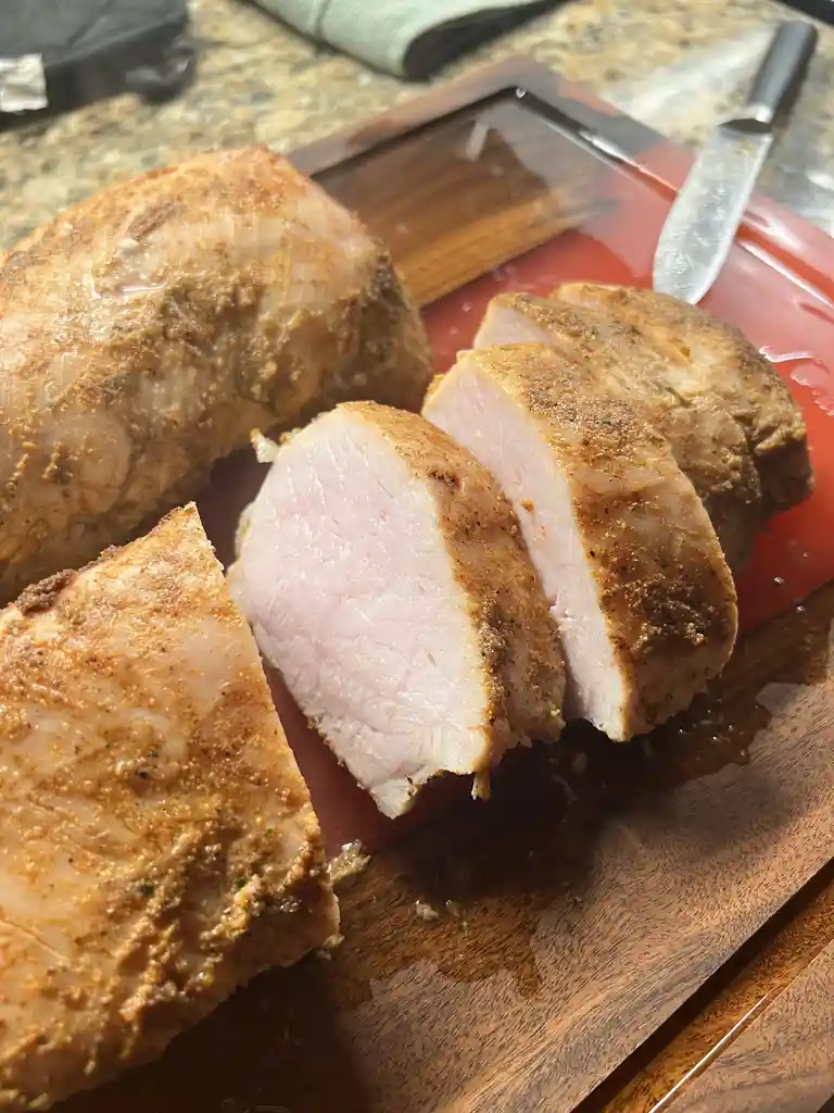 A close-up photo shows a cooked pork roast on a wooden cutting board with a red mat underneath. Several slices have been cut…