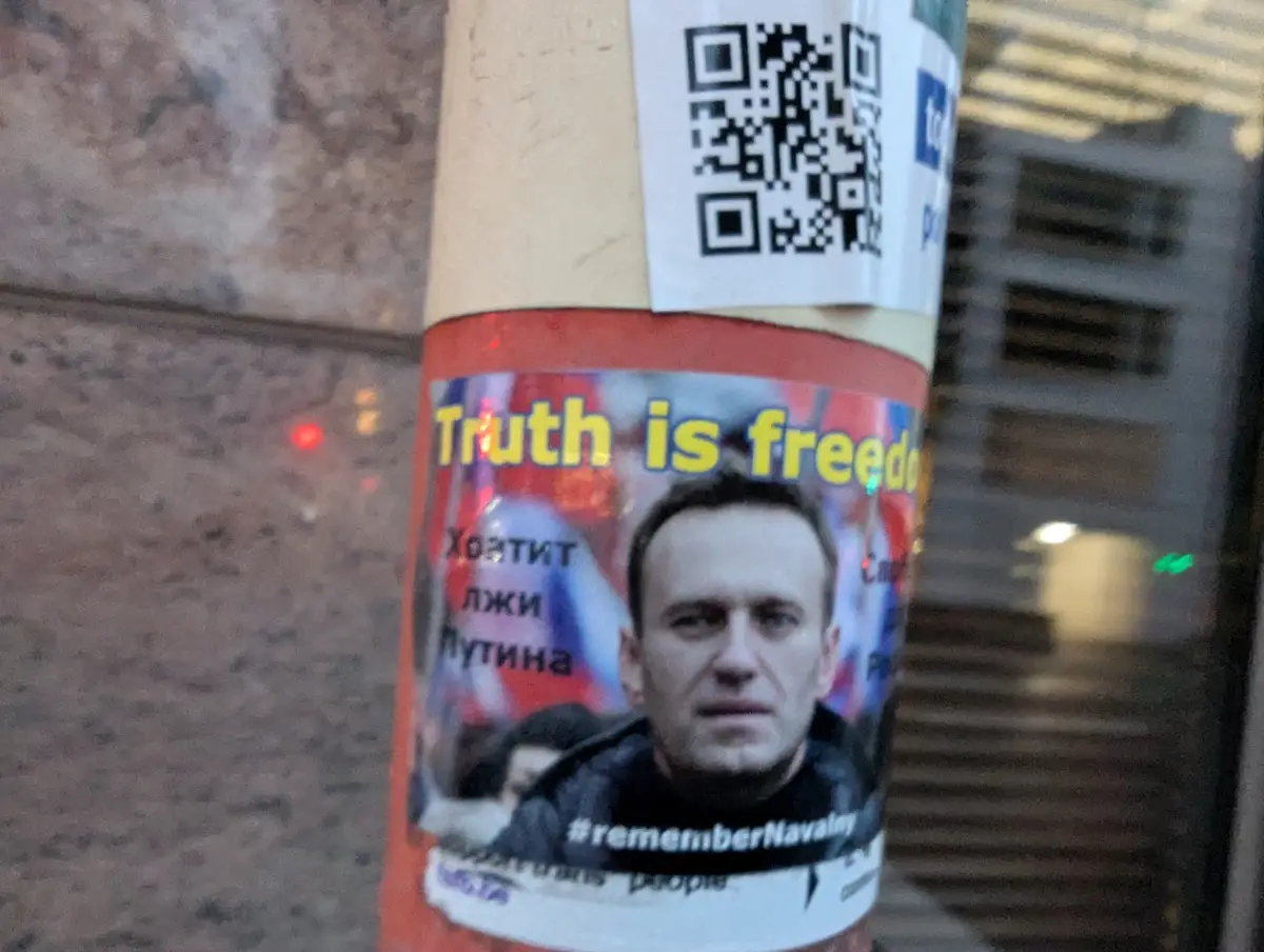 A close-up photo of a protest sticker on a traffic cone, featuring a photo of Alexei Navalny. The sticker has the English…
