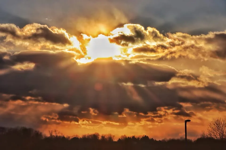 A dramatic sunset with golden sunbeams piercing through dark clouds, silhouetting trees and a streetlamp on the horizon. The…