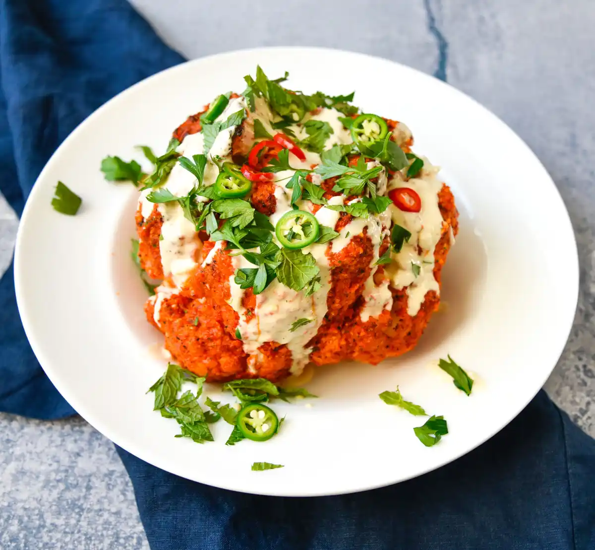 A white plate holds a large, orange-colored, textured food item resembling a cauliflower, topped with creamy white sauce…