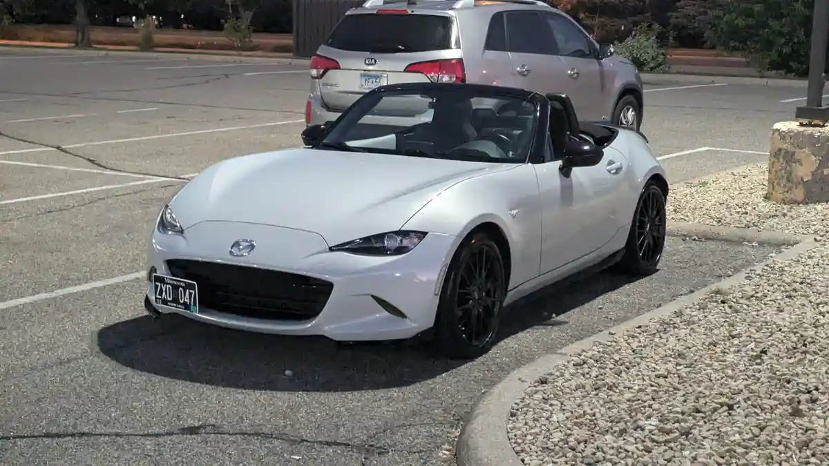 A white Mazda Miata convertible is parked in a lot at night, its top down, with a silver Kia Sorento behind it. The…