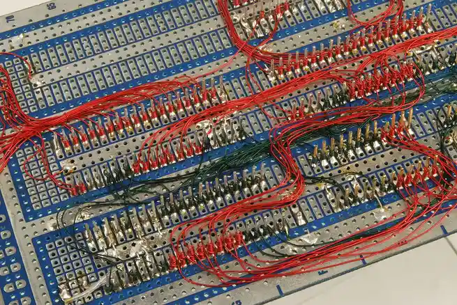 a close-up photo of a blue and silver breadboard densely populated with red and green jumper wires connected to its metal…