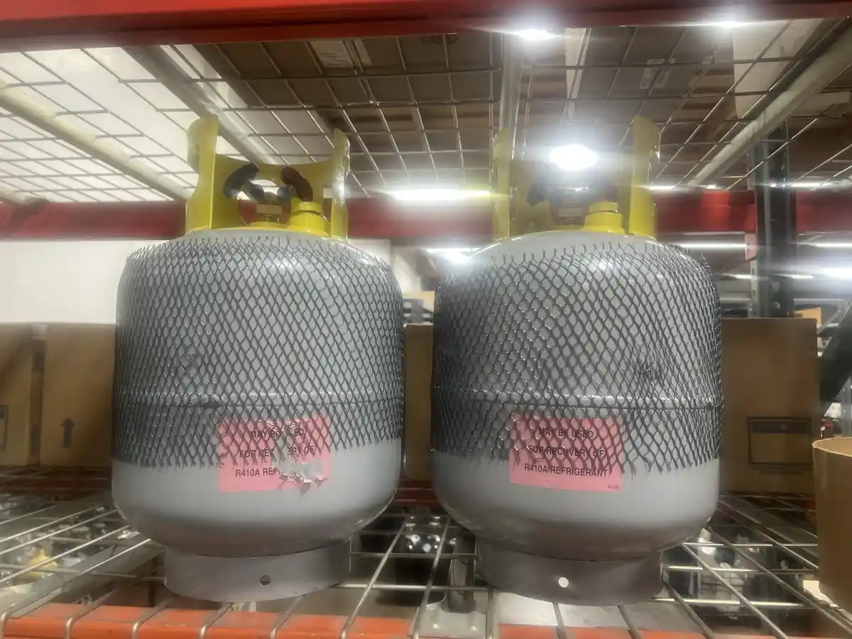 Two identical gray refrigerant cylinders with yellow valve caps sit side-by-side on a metal shelf in a warehouse. Each…